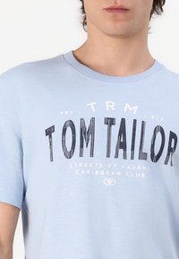 Gaišzils kokvilnas T-krekls ar apaļu apkakli, īsām piedurknēm un grafisku apdruku ar uzrakstu "TOM TAILOR" un tumši zilām strukturētām detaļām.