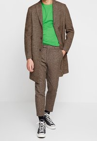 Brun houndstooth frakke, grøn t-shirt, brune strukturerede bukser og sorte sneakers med hvide såler. Afslappet sæt med kontrastfarver.