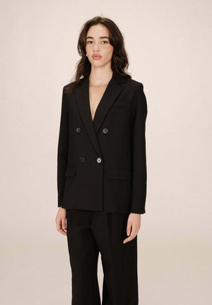 Femme aux longs cheveux noirs portant un blazer noir à double boutonnage et un pantalon assorti, devant un fond beige uni.
