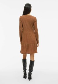 Frau steht mit dem Rücken zur Kamera, trägt ein braunes, geripptes Langarmkleid mit einem gestuften Rock und schwarze, kniehohe, Absatzstiefel.
