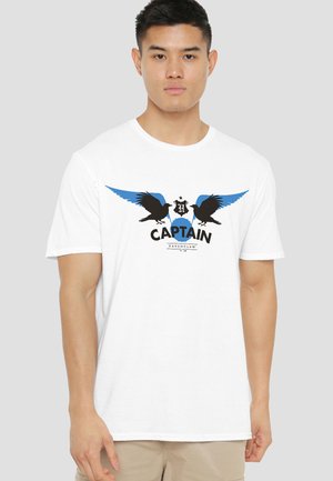 Young man wearing white t-shirt with black ravens, blue wings, Hogwarts crest, and text "Captain Ravenclaw" on front.