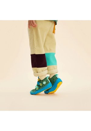 Enfant portant un pantalon beige bicolore et des baskets à scratch bleu-vert avec des semelles jaunes, se tenant sur la pointe des pieds devant un fond uni.