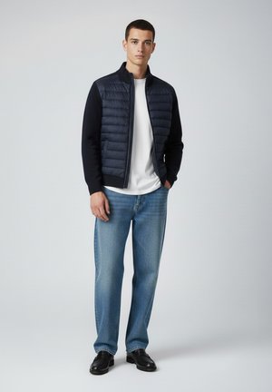 Młody mężczyzna stojący w granatowej pikowanej kurtce, białej koszulce, niebieskich dżinsach i czarnych skórzanych butach na neutralnym tle.