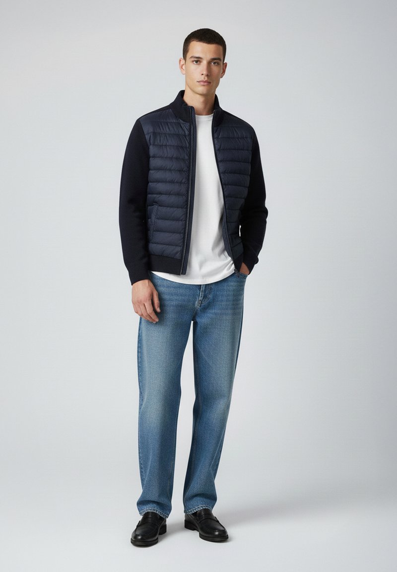 Giovane uomo in piedi che indossa una giacca trapuntata blu navy, una t-shirt bianca, jeans blu e scarpe di pelle nere su uno sfondo neutro.