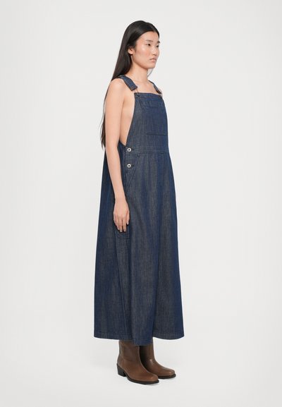Rochie din denim cu picioare late, bretele ajustabile și butoane pe lateral. Culoare albastru închis, până la genunchi, asortată cu cizme negre până la gleznă.