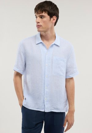 Ung mand med kort brunt hår iført en lyseblå, stribet kortærmet skjorte med knapper og mørkeblå bukser, ser mod venstre.