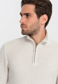 Un pull zippé beige clair présente un design en maille texturé avec un col côtelé et une courte fermeture éclair, offrant un look moderne et respirant.