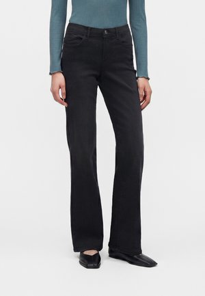 Femme portant un jean noir taille haute à jambes droites, un top côtelé à manches longues couleur sarcelle et des chaussures noires à bouts carrés pointus, debout sur fond blanc.