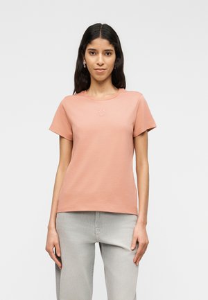 Hosszú sötét hajú nő egyszerű barackszínű rövid ujjú pólót és világosszürke nadrágot visel, egy egyszerű világos háttér előtt áll.