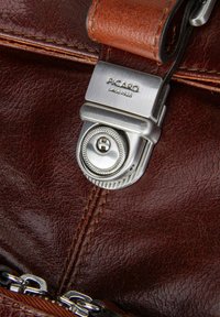 Leren tas sluiting in zilver met "PICARD sinds 1928" gegraveerd, bevestigd aan een bruine leren schouderband en met een gestikt leren oppervlak.