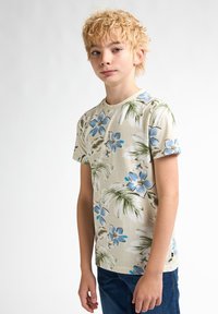Lysbeige skjorte med blå blomsterprint og grønne palmeblade. Korte ærmer, rund hals, lavet af blødt stof, kombineret med blå jeans.