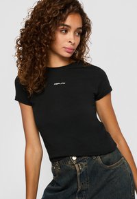 Jonge vrouw met krullend haar, gekleed in een zwart Replay t-shirt en donkerblauwe spijkerbroek, kijkt iets naar haar links.