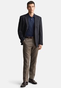 Námornícka polokošeľa pod pruhovaným sakom, doplnená hnedými textúrovanými nohavicami a tmavými topánkami. Sako má vertikálne pruhy a vrecko.