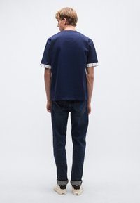 Tmavomodré tričko s krátkym rukávom a kontrastnými károvanými manžetami, skombinované s tmavomodrými džínsami a béžovými teniskami s čiernymi detailmi.