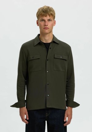 Jeune homme aux cheveux blonds courts portant une veste-chemise vert foncé boutonnée avec des poches à rabat, debout devant un fond clair uni.