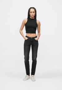 Čierny krátky top bez rukávov, tmavé rifle s vysokým pásom, biele tenisky, stojaci s rukami v vreckách, vzpriamená postura na jednoduchom pozadí.