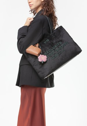 Borsa tote in velluto nero con testo ricamato verde, dotata di una pochette in pelle color nocciola e un portachiavi floreale. Modello che indossa un blazer nero e pantaloni color ruggine.