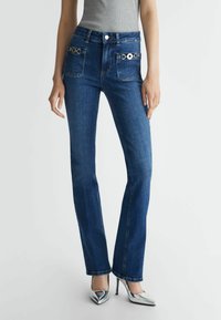 Donkerblauwe flared jeans met twee zijzakken, zilveren hardware accenten en een gladde denim textuur, gecombineerd met zilveren hoge hakken.