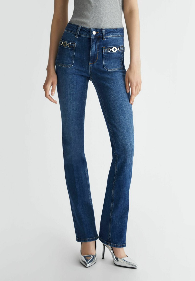 Donkerblauwe flared jeans met twee zijzakken, zilveren hardware accenten en een gladde denim textuur, gecombineerd met zilveren hoge hakken.