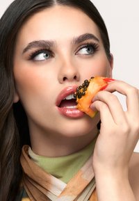 Zbliżenie osoby trzymającej plaster papai z czarnymi nasionami na wierzchu, ubraną w wzorzysty szalik. Usta są błyszczące i lekko rozchylone.