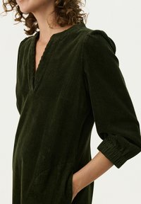 Dunkelgrünes Cordkleid mit V-Ausschnitt, Dreiviertelärmeln und seitlichen Taschen. Hat eine gerippte Struktur und lockere Passform.