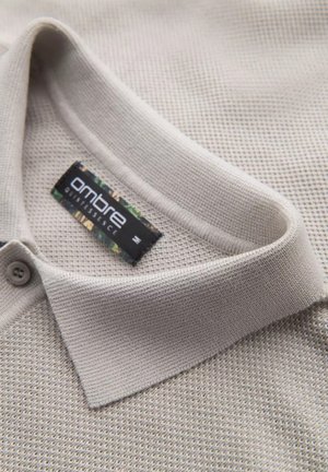 Polo shirt beige chiaro in tessuto testurizzato, con scollatura a colletto e patta con due bottoni. L'etichetta mostra il marchio "ombre".