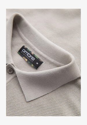 Polo de color beige claro con una textura suave, con cuello y cierre de dos botones. La etiqueta muestra la marca "ombre".