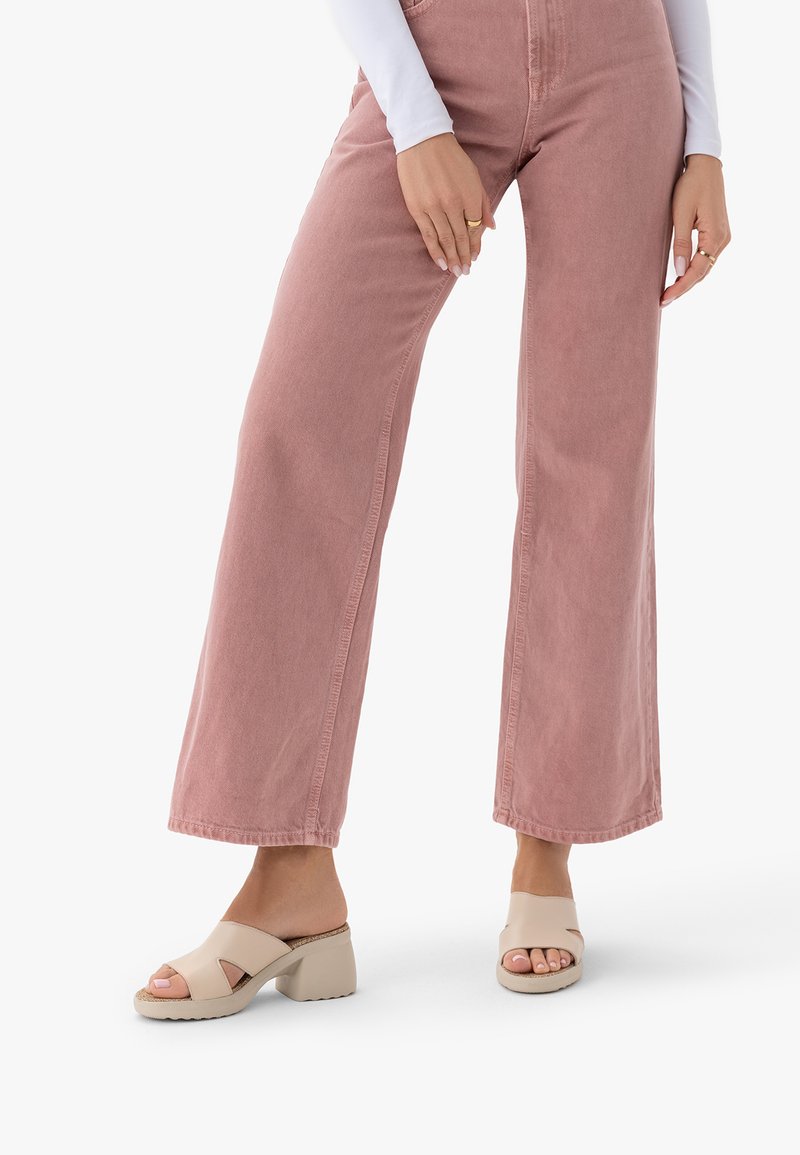 Nő rózsaszín, bő szárú nadrágot és bézs, nyitott orrú sarkú szandált visel, fehér háttér előtt áll.