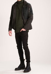 Chaqueta negra acolchada con un bolsillo con cremallera, suéter verde oliva, jeans negros y botas negras con cordones. Ajuste moderno y diseño en capas.