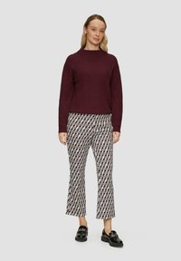 Un pull côtelé bordeaux à col montant, assorti à un pantalon évasé à motifs blancs, rouges et noirs. Des mocassins noirs brillants complètent le look.