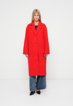 Langer roter Wollmantel mit Reverskragen, zwei Fronttaschen und einer Knopfleiste mit zwei Knöpfen. Trägt man über weiten blauen Jeans und schwarzen Schuhen.