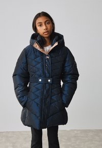 Marineblauwe doorgestikte donsjas met capuchon, getailleerde taille en contrasterende roségouden voering, voorzien van drukknopaccenten en diagonale stiksels.