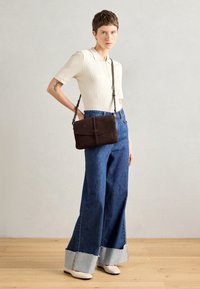 Borsa a tracolla in suede marrone scuro con dettagli intrecciati e nappina; indossata con un polo leggero a coste e pantaloni di jeans a gamba larga con orlo grezzo.
