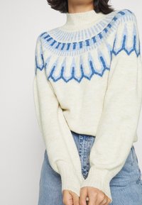 Woman wearing a cream sweater with blue patterned yoke and high neckline, paired with light blue high-waisted jeans.