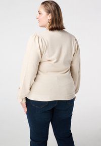 Roomkleurige trui met opgeblazen mouwen, geribbelde manchetten en een nauwsluitend ontwerp, gecombineerd met donkerblauwe jeans. Gladde textuur en klassieke vorm.