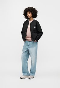 Giacca nera trapuntata con tasca applicata, felpa rosa chiaro con scritta bianca, jeans oversize azzurro chiaro e sneakers grigie.