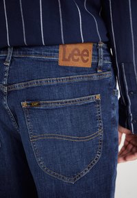 Ciemnoniebieskie dżinsy z denimu z brązową skórzaną metką oznaczoną napisem "Lee" na pasku. Posiadają żółte przeszycia oraz standardowy wzór tylnej kieszeni.