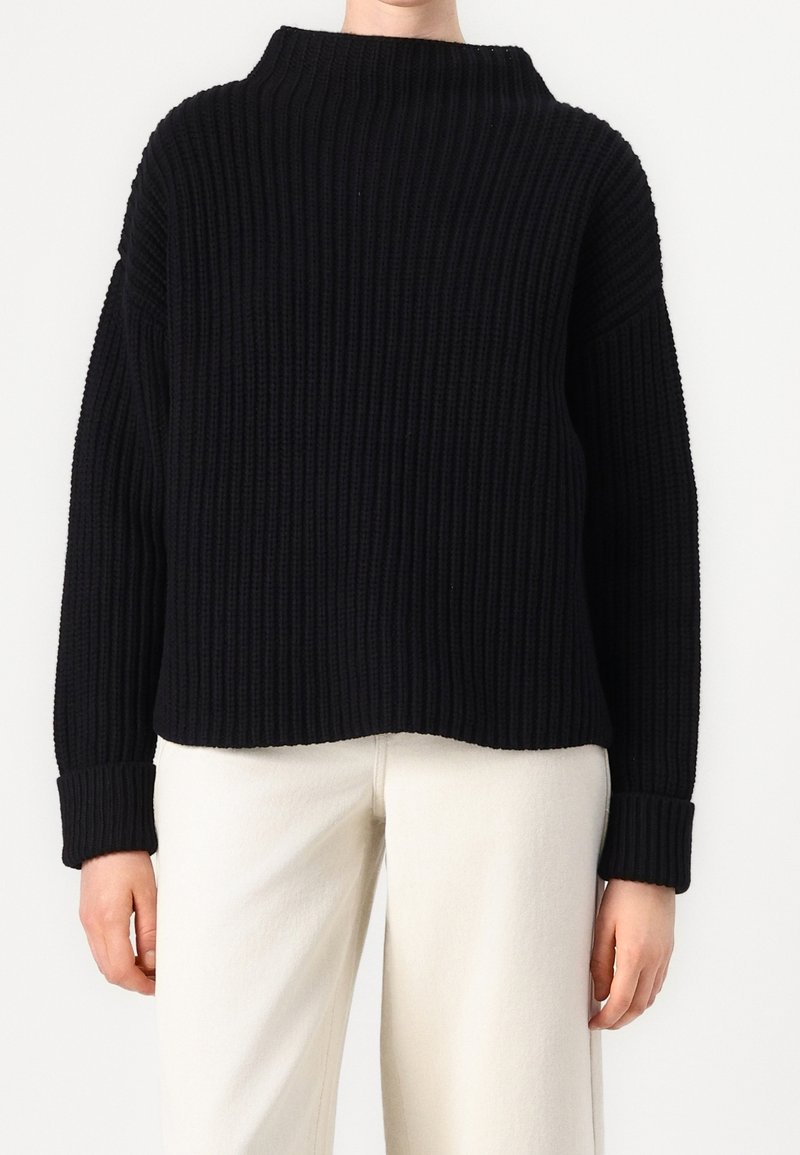 Pull noir côtelé avec col haut et manches larges. Texture épaisse, coupe ample et poignets retroussés. Associé à un pantalon de couleur claire.