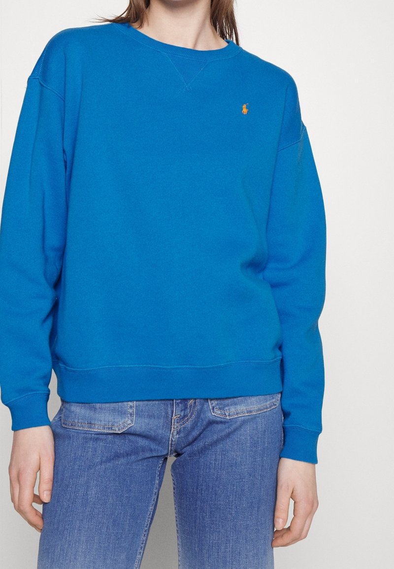 Blå bomulls sweatshirt med rund hals och ribbade ärmslut, med en broderad logga i orange. Matchad med blå denimjeans.