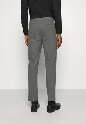 Pantalon gris ajusté avec une texture lisse, coupe slim et plis subtils. Porté avec des chaussures noires et une chemise noire à manches longues.