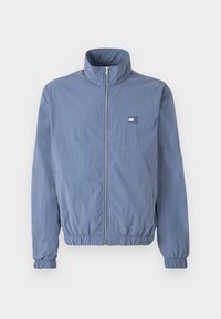 Veste bleu clair zippée avec col montant, poignets et ourlet élastiques, et petite étiquette rectangulaire avec logo sur la poitrine gauche.