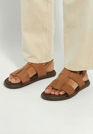 Brown leather sandals with crisscross straps, flat soles, and open toe design. Worn with light-colored, cuffed pants.