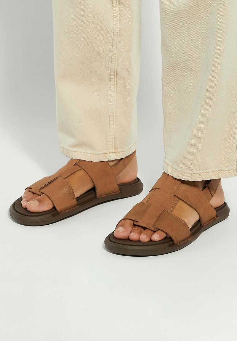 Brown leather sandals with crisscross straps, flat soles, and open toe design. Worn with light-colored, cuffed pants.