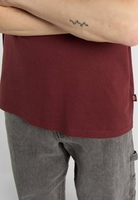 Burgundská bavlněná trička s kulatým výstřihem spárovaná se šedými texturovanými džíny s cargo kapsami a poutky na opasek. Viditelné jemné detaily v prošívání.