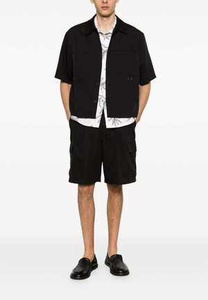 Uomo che indossa una camicia nera a maniche corte con bottoni, una camicia floreale bianca sotto, pantaloncini cargo neri e scarpe slip-on nere, in piedi con le mani nelle tasche.