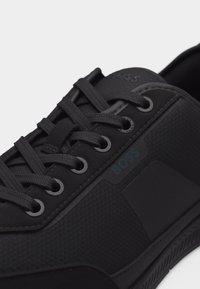 Μαύρο αθλητικό παπούτσι χαμηλού ύψους με υφή στις πλευρές, μαύρα κορδόνια, μεταλλικές οπές και λογότυπο "BOSS" κοντά στα κορδόνια σε λευκό φόντο.