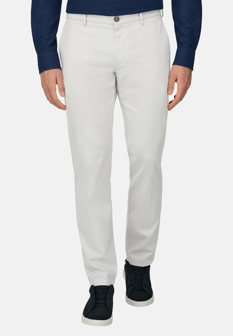 Homme portant un pantalon blanc ajusté, une chemise bleu marine à manches longues et des baskets noires avec semelles blanches, debout devant un fond uni.