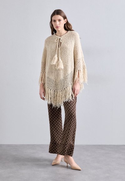 Strikket poncho i krem med frynset kant og dekorative dusker, paret med brune mønstrede slengbukser og nude hæler.