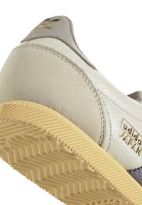 Grå och beige lädersneakers med texturerad yta, visar en sula av gummigummi med distinkt mönster och detaljer i guld.