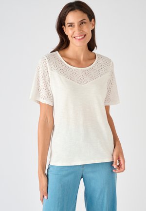 Glunderende vrouw die een wit kanten top met korte mouwen en lichtblauwe broek draagt, staand tegen een effen lichte achtergrond.